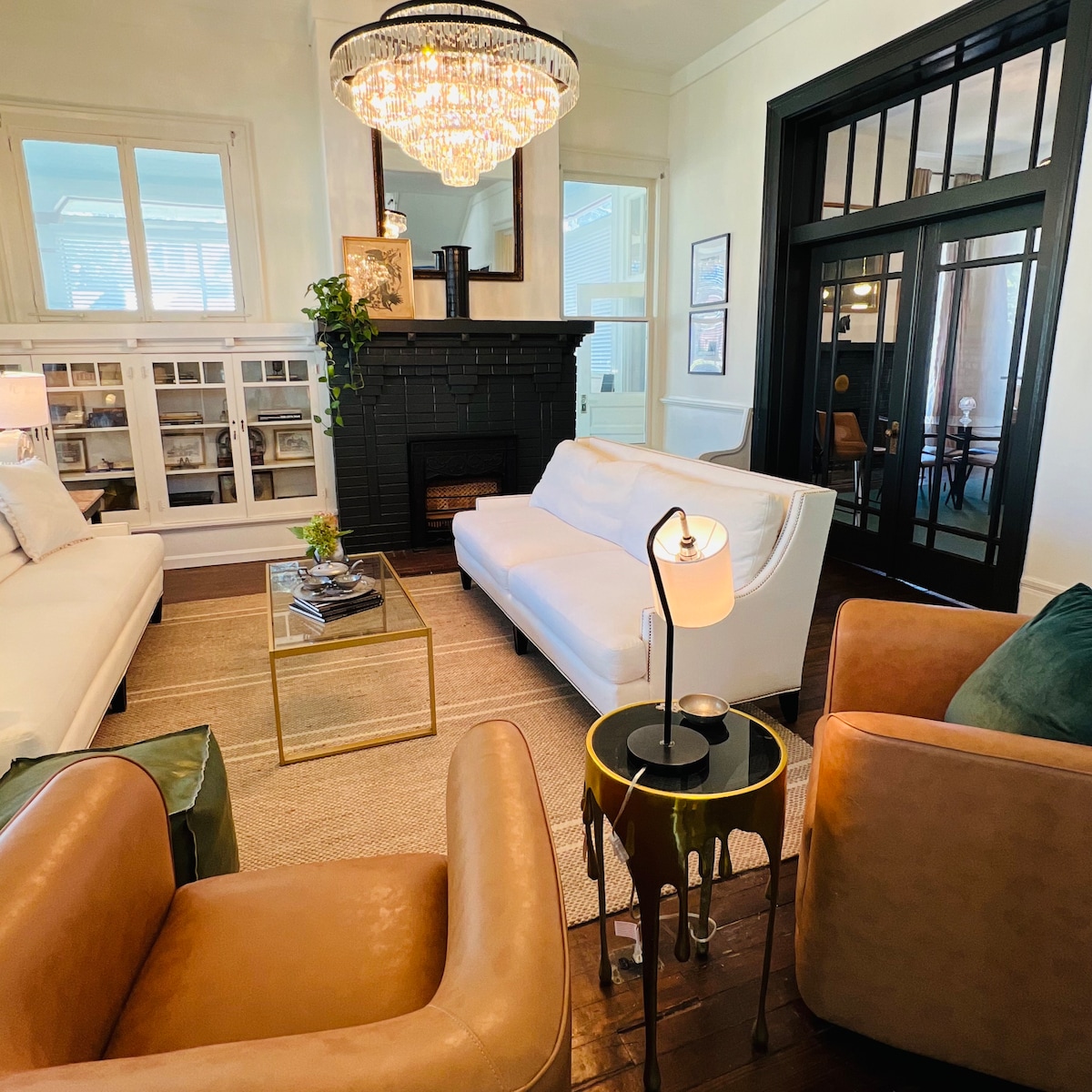 A stylish living area features a modern chandelier above a glass coffee table, surrounded by white sofas and brown armchairs. A black fireplace adds a classic touch, while large windows allow natural light to brighten the space. A potted plant adds a hint of greenery.
