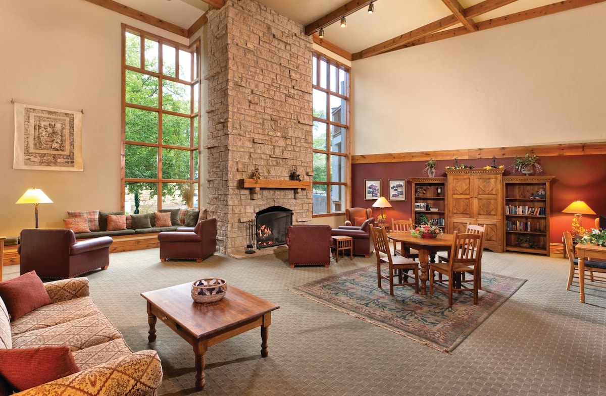 A spacious common area is highlighted by a tall stone fireplace at the center. Large windows allow natural light to fill the room, complemented by wooden beams overhead. Comfortable seating arrangements include leather chairs and wooden tables, creating a welcoming space for relaxation.