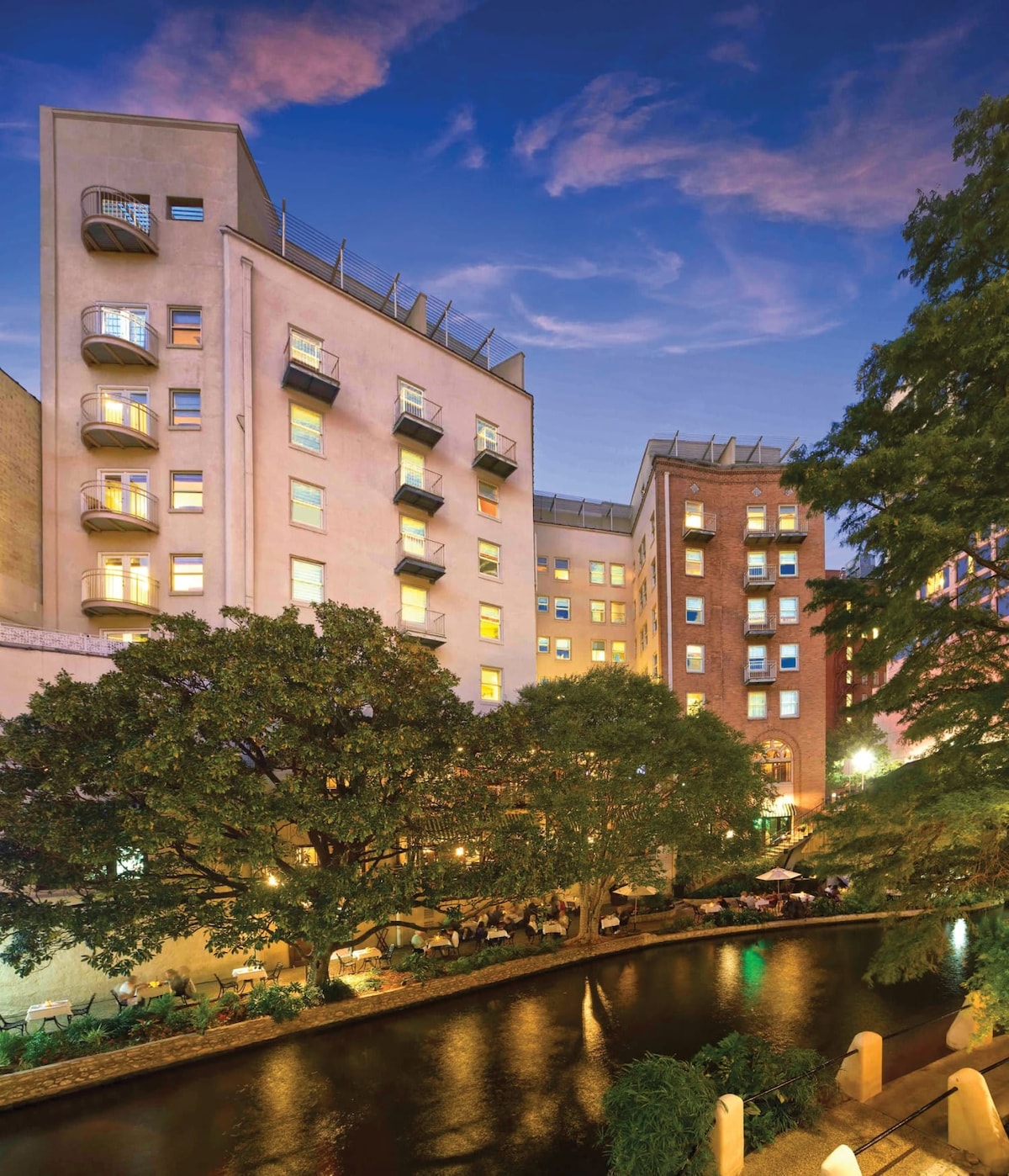 The view captures the historic resort building alongside the River Walk at dusk, with soft lighting emanating from the windows. Balconies protrude from the structure, while trees and ambient lighting provide a serene atmosphere along the water's edge.