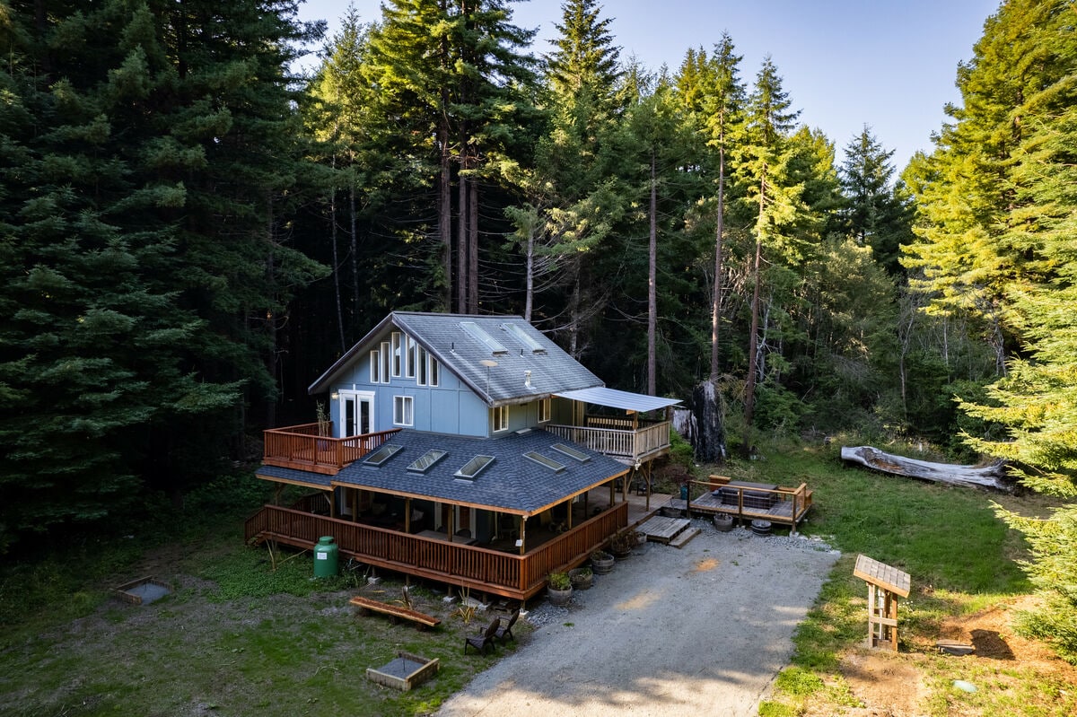 The retreat is surrounded by towering trees, featuring multiple outdoor decks that offer spacious areas for relaxation. A gravel pathway leads to the entrance, where natural light filters through the forest canopy, creating a serene atmosphere in this private setting.
