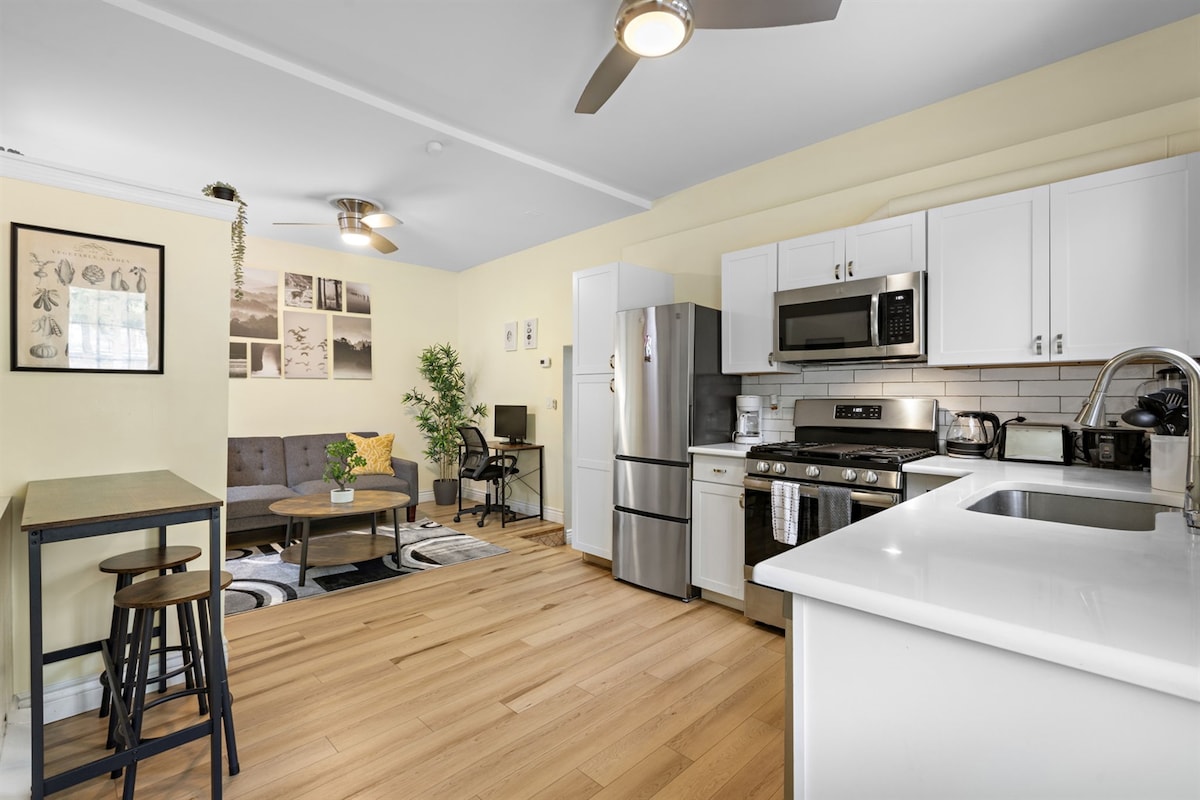 The kitchen area features modern stainless steel appliances, including a refrigerator and gas stove. A dining table with stools is positioned beside a cozy seating area with a sofa and decorative plants. Natural light filters in through the window, illuminating the warm wood flooring.