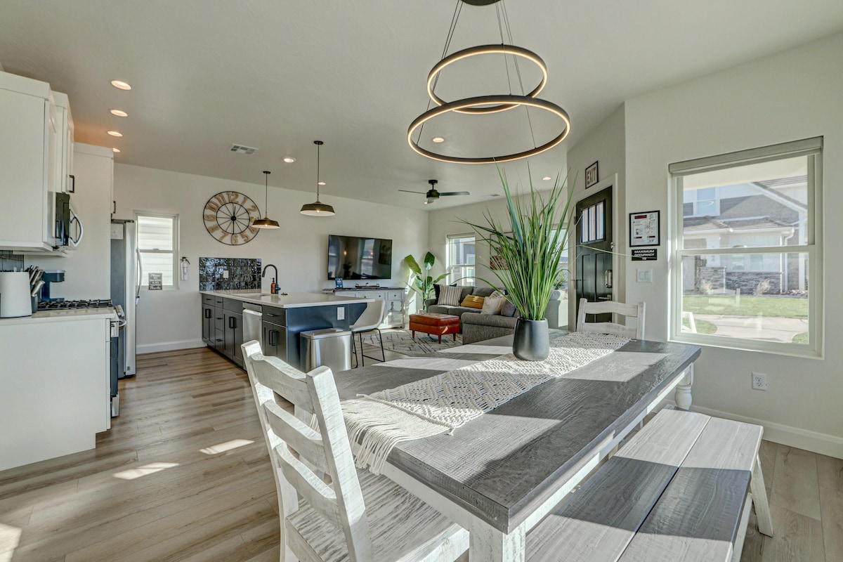 An open-concept living area features modern furnishings, including a spacious dining table with light-colored chairs. A mix of natural light and warm overhead lighting enhances the inviting space. A comfortable sofa and television are positioned for relaxation, while tasteful decor elements and plants add a touch of style.