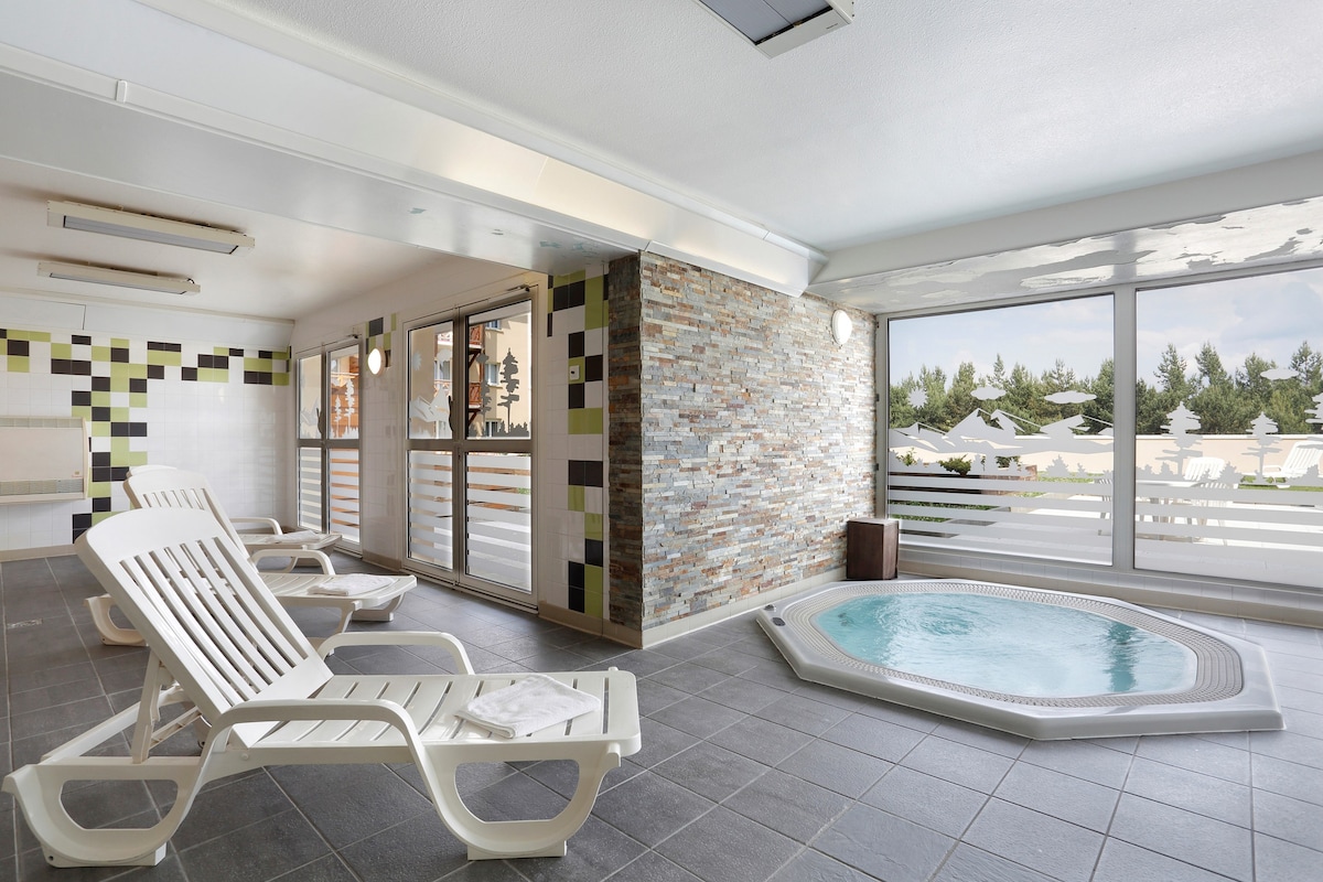 A bright indoor wellness area features a hot tub surrounded by tiled walls and large windows offering views of the outdoors. Several lounge chairs are positioned along one side, inviting relaxation in a serene environment.