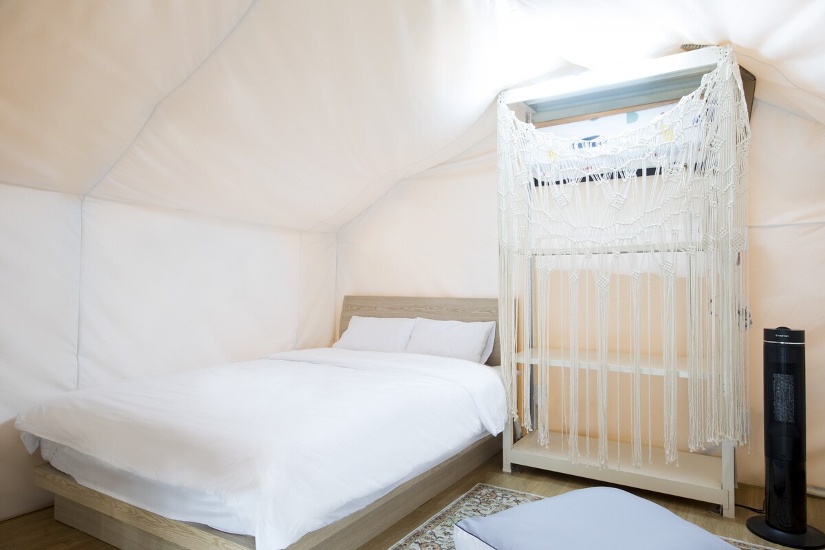 A bright sleeping area features a spacious bed dressed in crisp white linens. A decorative macramé wall hanging is mounted above a wooden shelf, which stands adjacent to a modern standing fan. Soft natural light from a window enhances the serene atmosphere.