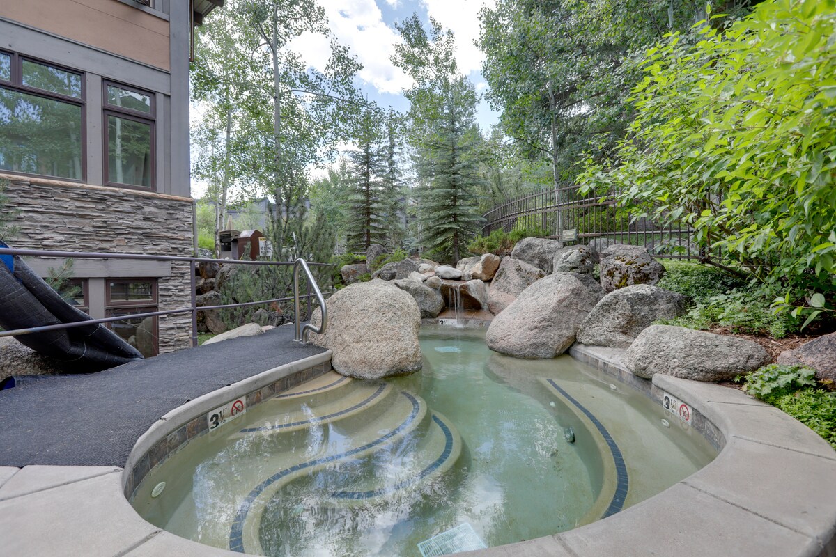 A communal hot tub is surrounded by natural stone and lush greenery. Smooth rocks and boulders create a rustic ambiance, while the serene water features hint at a gentle waterfall nearby. An inviting path leads to the hot tub area, offering a peaceful retreat.