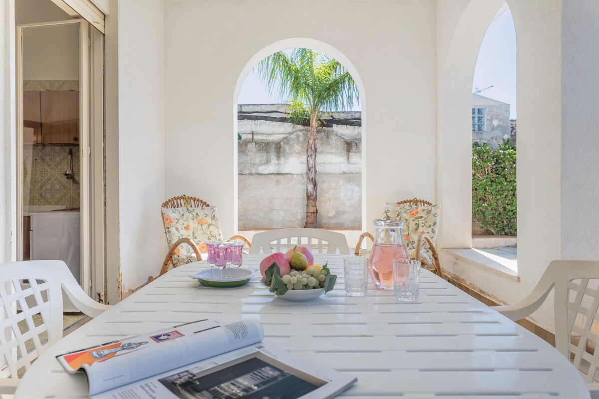 A spacious outdoor terrace features a large dining table surrounded by white chairs. A tropical palm is visible through an arched window, and a bowl of fresh fruits is placed at the center of the table. Two comfortable chairs add to the inviting setting.
