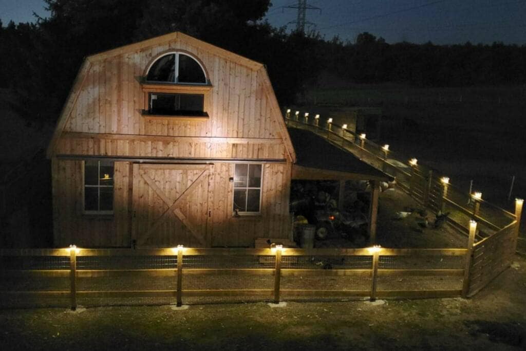 The exterior of the barn is illuminated at night, revealing wooden paneling and large windows. A gentle glow is provided by lights along the fenced pathway that leads to the entrance. The surrounding rural landscape is visible in the background.