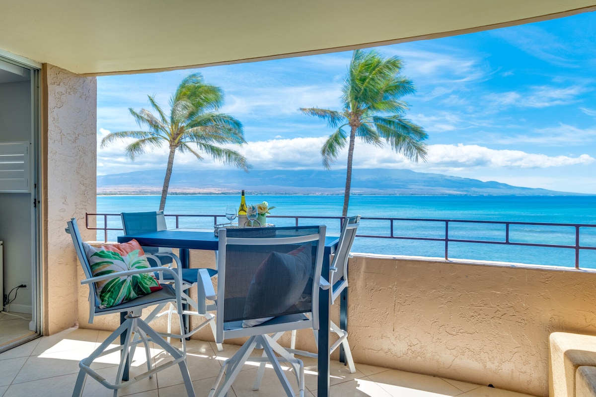 A spacious outdoor lanai features a dining table with four chairs, positioned to take in panoramic ocean views. Lush palm trees sway gently in the foreground, while the clear blue water and distant mountains create a serene backdrop.