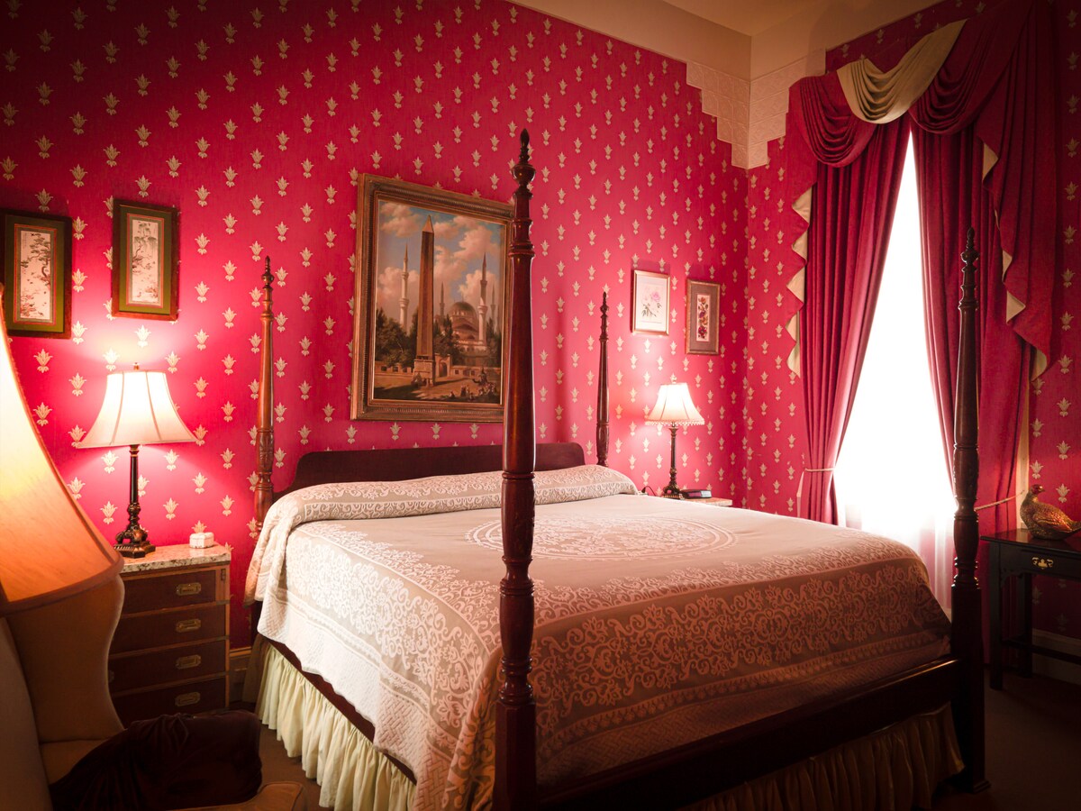 A large room features bold red floral wallpaper and elegant drapery framing a bright window. A wooden poster bed is adorned with decorative bedding. Soft lighting from bedside lamps creates a warm ambiance, while a vintage painting adds a classical touch to the decor.