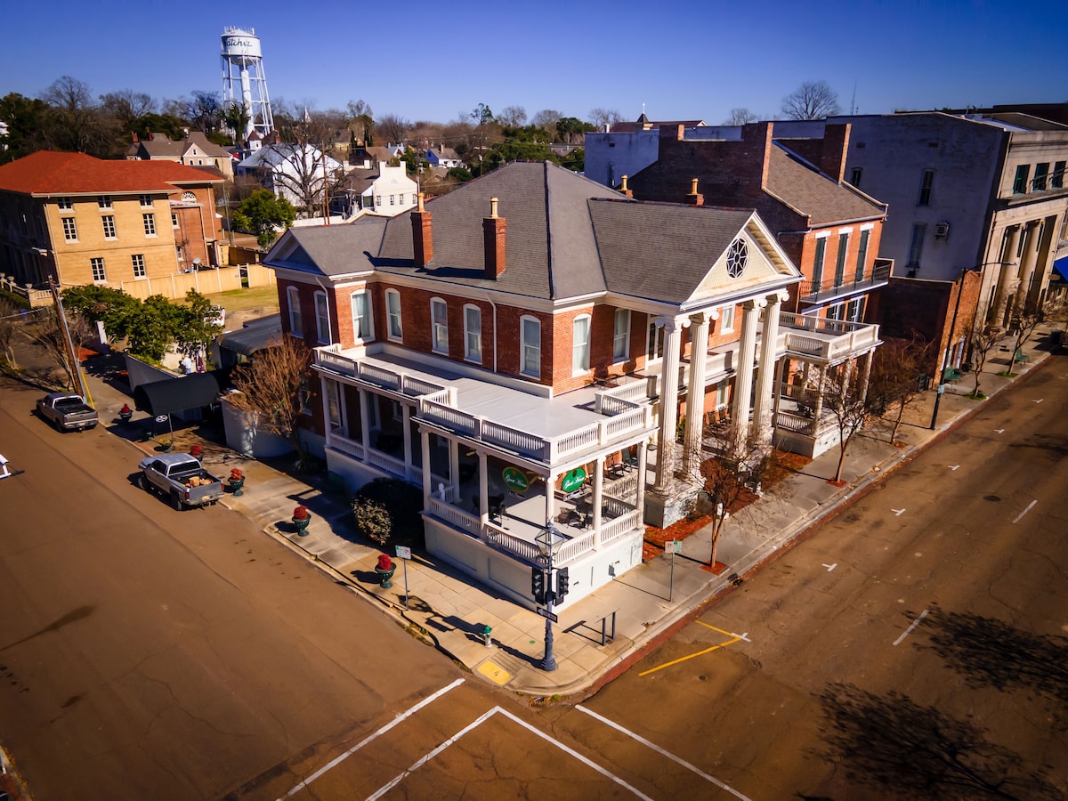 A historic building is viewed from above, showcasing a large brick structure with white columns. A covered porch runs along the front, and several trees are visible near the street. The surrounding area includes several vehicles and a water tower in the background.