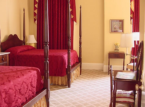 A spacious room features two four-poster beds dressed in rich burgundy linens. Light yellow walls enhance the bright atmosphere, while red drapes frame the windows. A small writing desk and chair are positioned near the entrance, adding functional elegance to the space.