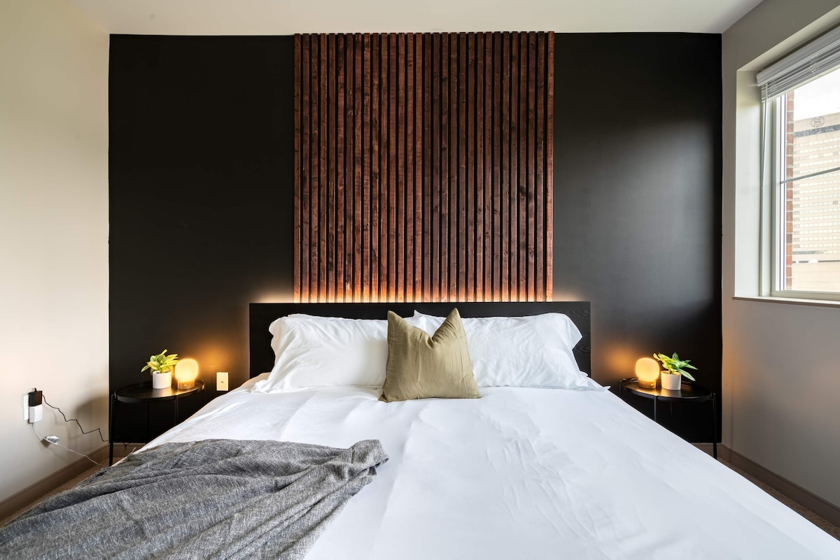 The bedroom features a spacious bed with crisp white linens and a decorative throw blanket, positioned against a dark wall accented by wooden paneling. Nightstands with gentle lighting are situated on either side, while a window allows natural light to enrich the ambiance.