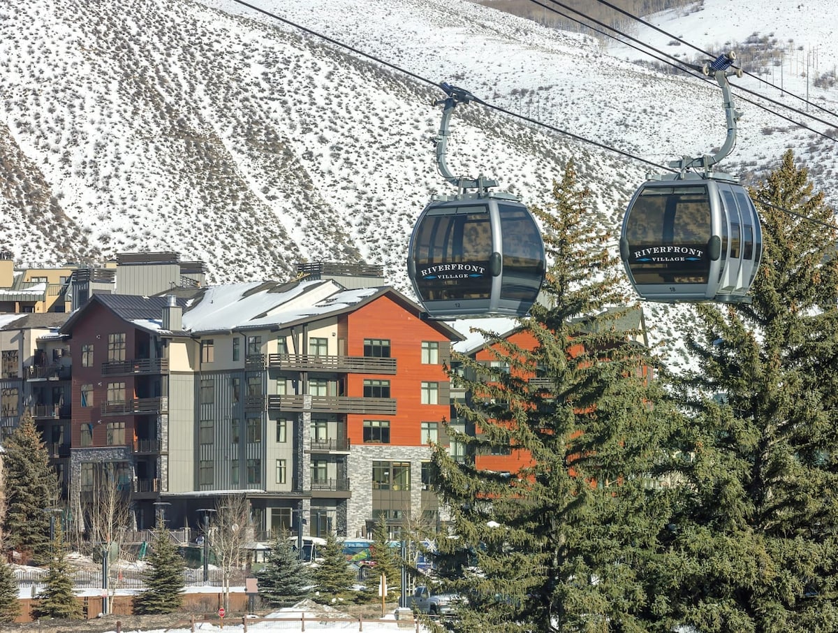 A scenic view of a gondola transport system, with two cabins gliding above a snow-covered landscape. The backdrop features a modern building with varying colors, surrounded by tall evergreen trees. Rocky mountains rise in the distance, providing a striking natural setting.