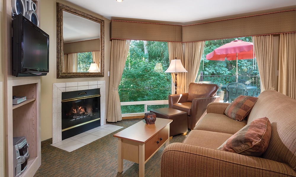 A cozy living area features a comfortable sofa and armchair, with a flat-screen television mounted on the wall. A fireplace is situated beneath a large mirror, and large windows offer views of lush greenery. A patio with an umbrella is visible through the windows.