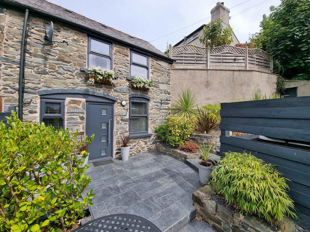 The image shows a charming stone cottage with a slate patio area. Lush greenery and potted plants frame the entrance, enhancing the inviting atmosphere. The door is flanked by flower boxes, contributing to the cozy exterior. A wooden fence provides privacy along the side.