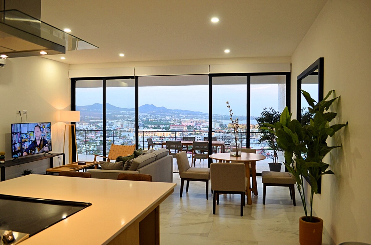 A spacious living area features large floor-to-ceiling windows, showcasing views of the city and mountains. A modern kitchen with an island connects to a comfortable seating area, which includes a sofa and armchairs. A dining table for four is positioned near the windows, creating a bright atmosphere.