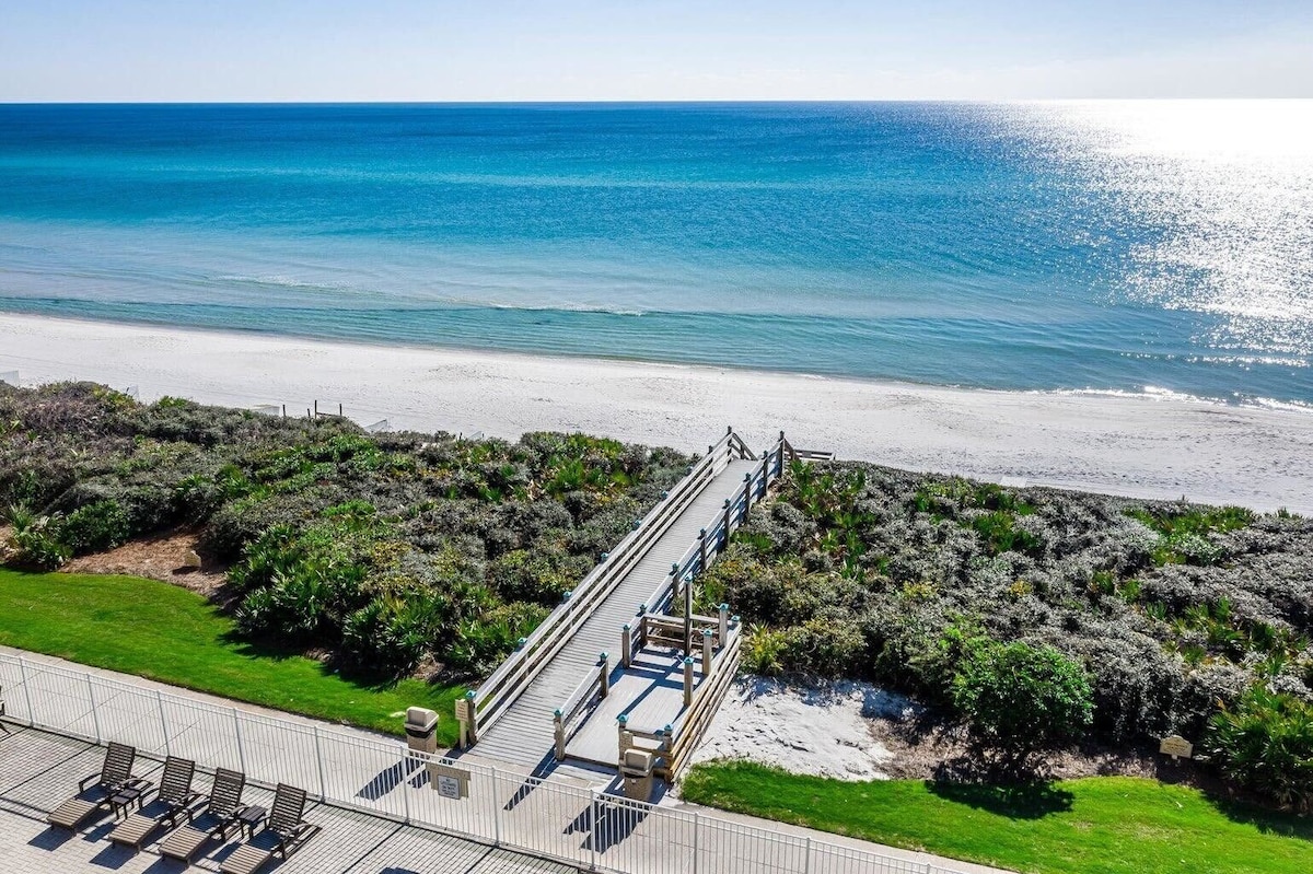 A wooden boardwalk leads from lush greenery to a private sandy beach, with gentle waves meeting the shore. The sun glistens on the clear turquoise water under a bright blue sky, creating a serene coastal atmosphere.