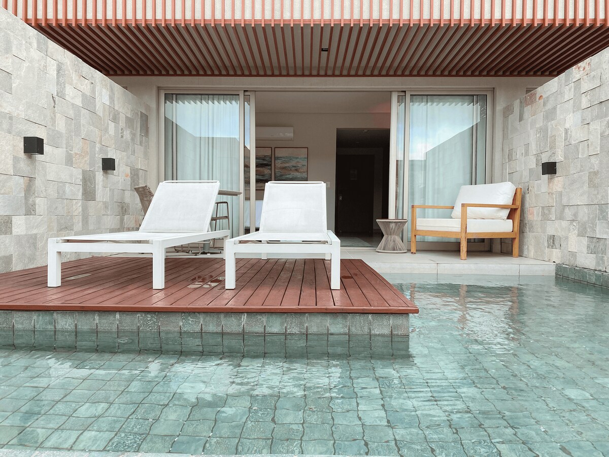 A deck with a wood surface is shown, featuring two white lounge chairs positioned beside a tranquil pool. Natural stone walls and large glass doors create a seamless transition to the indoor space, inviting relaxation in a serene environment.