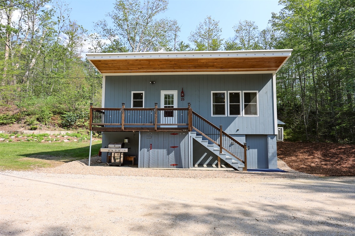 The exterior of the bungalow features a light blue facade with a wooden overhang and a front porch. Stairs lead to the entrance, and a gravel pathway is visible. Surrounding greenery enhances the home's outdoor setting, providing a natural and inviting atmosphere.