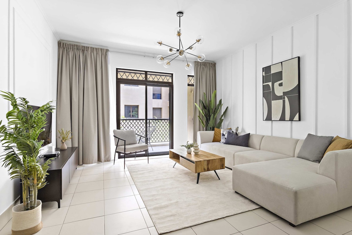 A spacious living area features a light-colored sectional sofa and a wooden coffee table atop a large area rug. Floor-to-ceiling curtains frame the windows, allowing natural light to fill the room. A television is placed on a low console, accompanied by decorative plants.