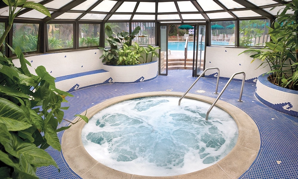 An indoor hot tub is surrounded by lush greenery and blue mosaic tiles. Natural light filters through the glass canopy, providing a warm ambiance. The area features two steps leading into the hot tub, enhancing accessibility.