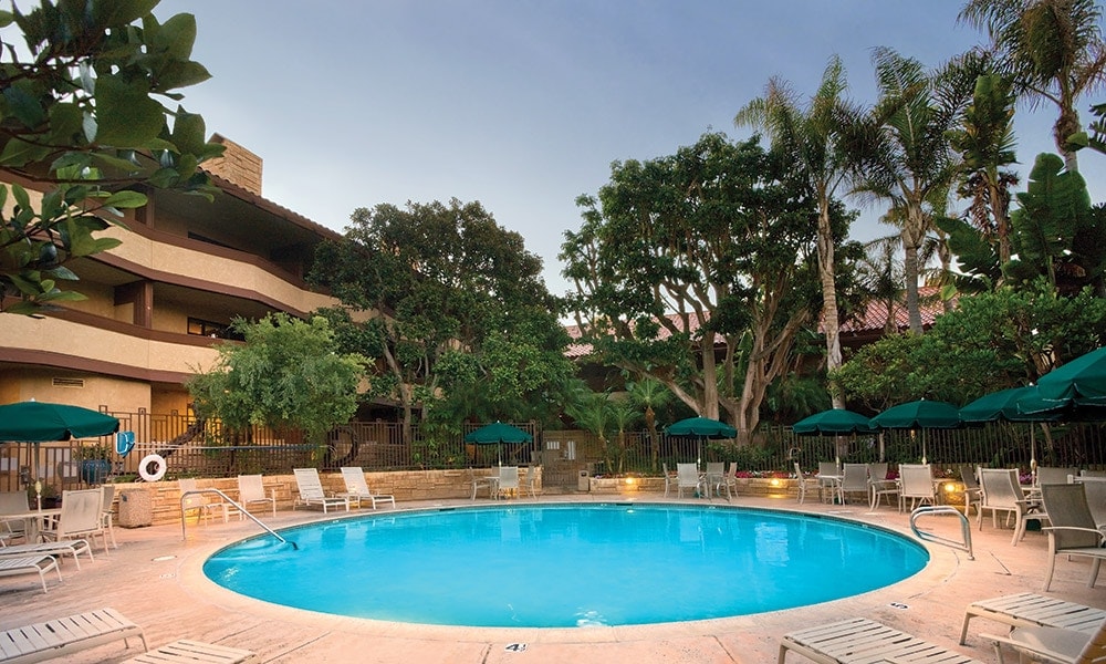 An outdoor swimming pool is surrounded by lush greenery and tropical trees, with several lounge chairs arranged along its edge. Umbrellas provide shade over some seating areas, while warm lights create a welcoming ambiance as evening approaches.