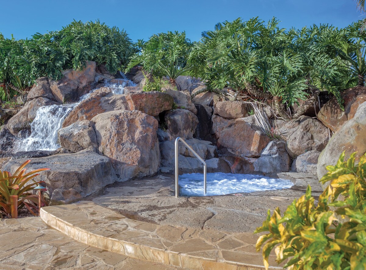 A serene outdoor area features a soothing hot tub surrounded by natural stone. A gentle waterfall cascades over rocks nearby, while lush greenery adds a tranquil ambience to the space.
