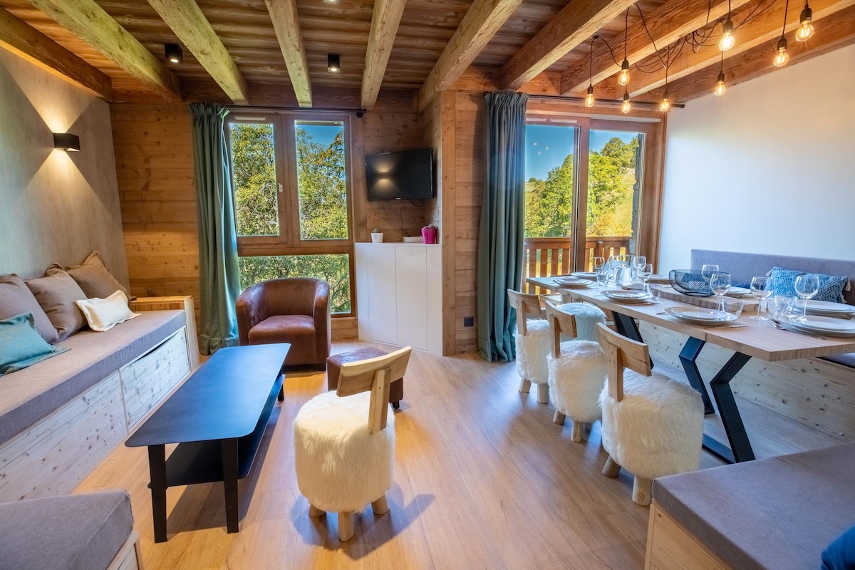A modern living area features large windows allowing natural light and showcasing views of the surrounding landscape. Comfortable seating includes a plush sofa and a brown armchair. A dining table is set with elegant glassware, complementing the warm wooden accents throughout the space.