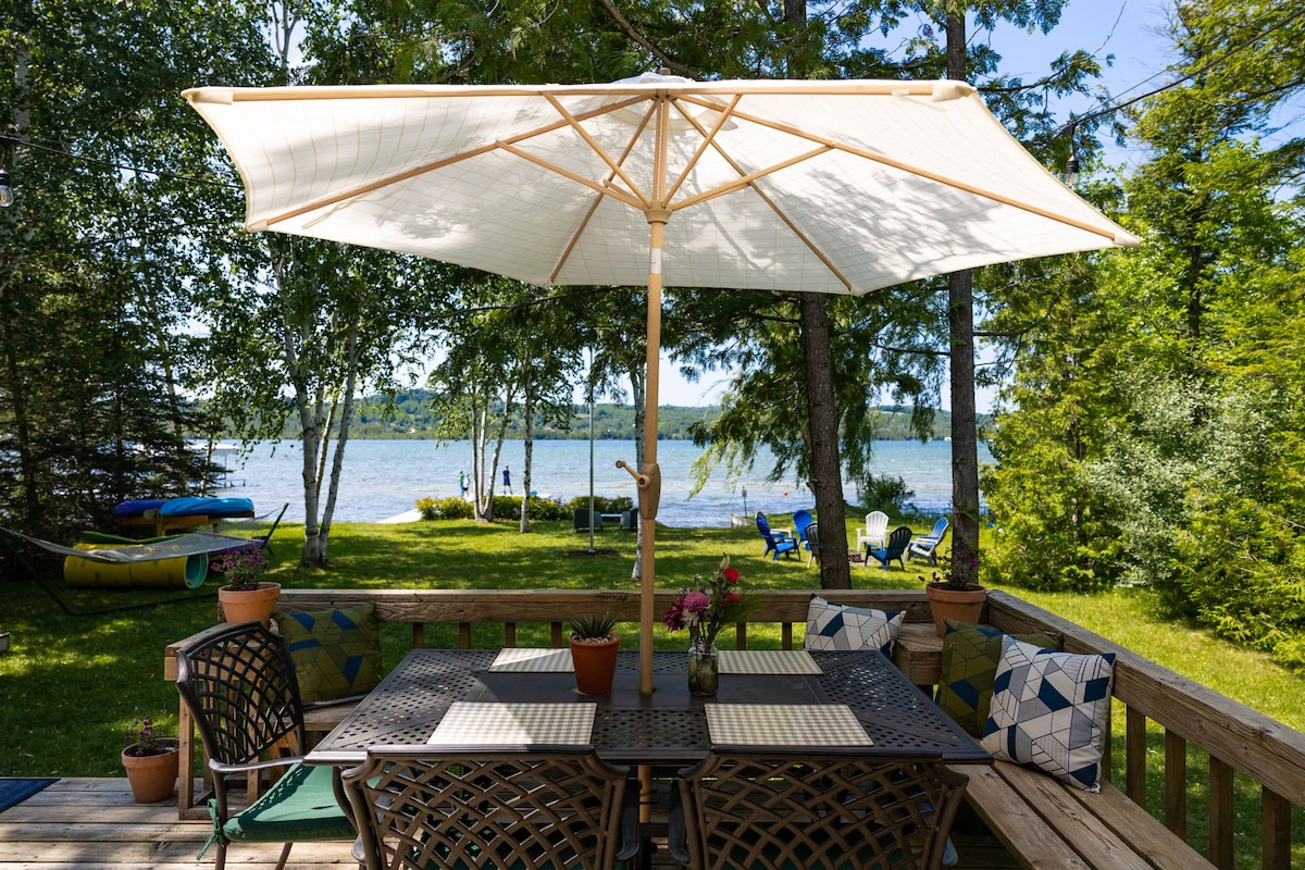 A wooden deck provides a shaded dining area with a large umbrella, surrounded by comfortable seating. Potted plants display vivid flowers, while the lush green yard leads to the sparkling lake visible in the background. Kayaks can be seen near the water's edge.