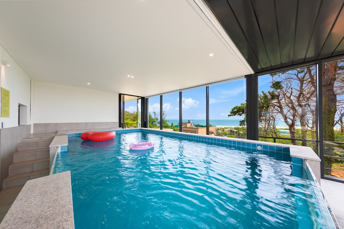An indoor pool is shown, featuring clear blue water and surrounded by large glass windows, offering views of the ocean and surrounding greenery. Colorful floating rings are placed in the water, adding a playful touch to the serene environment.