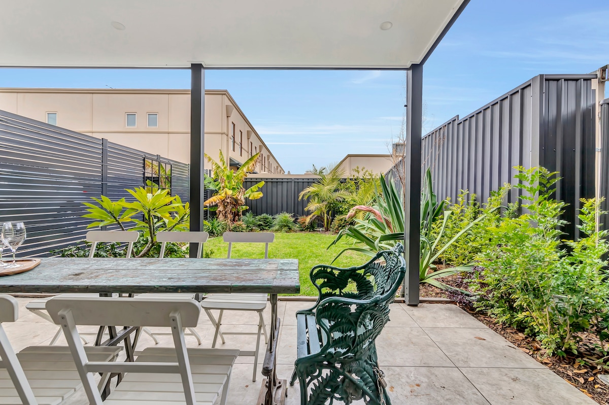 An outdoor dining area features a rustic wooden table and white chairs, surrounded by lush greenery. The garden is visible beyond the patio, showcasing vibrant plants and neatly trimmed grass, all framed by modern fencing that provides privacy.