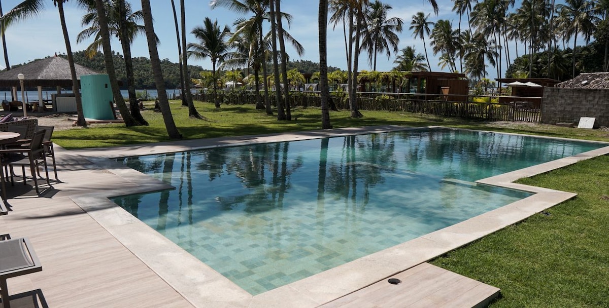 A spacious swimming pool is surrounded by lush green grass and tall palm trees. The water reflects the clear blue sky, providing a serene setting. Patio furniture is visible nearby, offering a place to relax and enjoy the tranquil landscape.