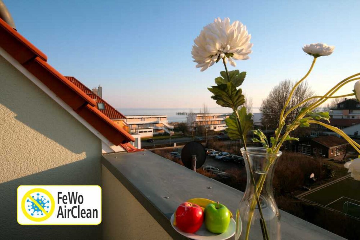 A balcony scene presents a clear view of the sea and surrounding area. A glass vase holding white flowers sits beside a bowl with red and green apples. The setting offers a peaceful atmosphere with a hint of the coastal landscape.