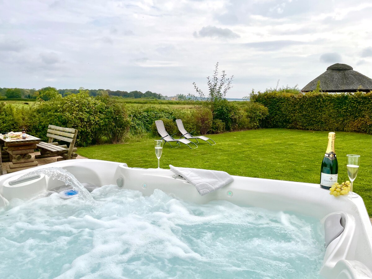 An outdoor whirlpool is filled with bubbling water, accompanied by two champagne flutes and a bottle of sparkling wine on the side. Green lawn stretches beyond towards a serene landscape, with two lounge chairs positioned for relaxation. A dining area set on a wooden table can be seen in the background.