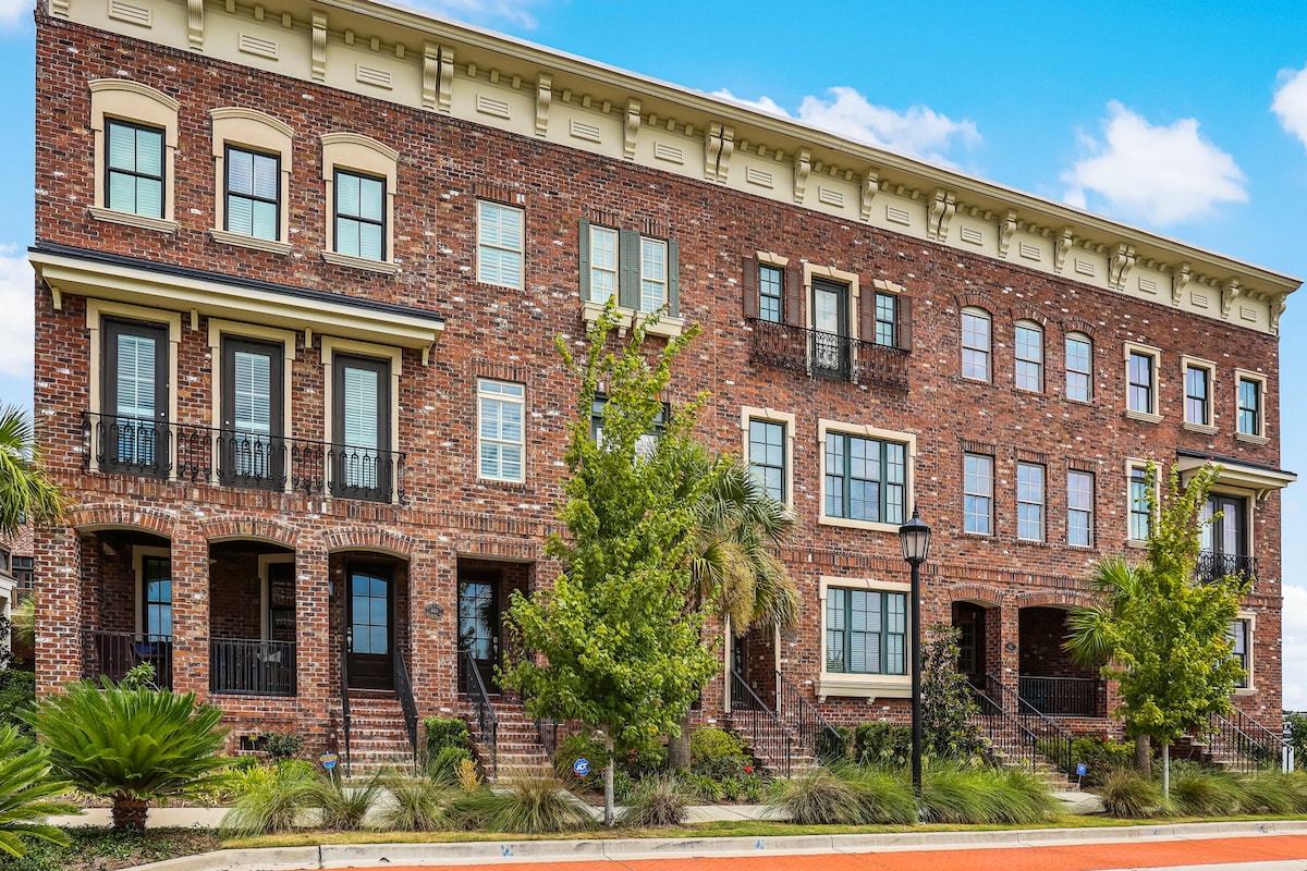 The exterior of a brick multi-level building is shown against a bright blue sky. Lush greenery lines the walkway leading to the entrance, and balconies with wrought iron railings add character. Large windows permit ample natural light, emphasizing the structure's inviting design.