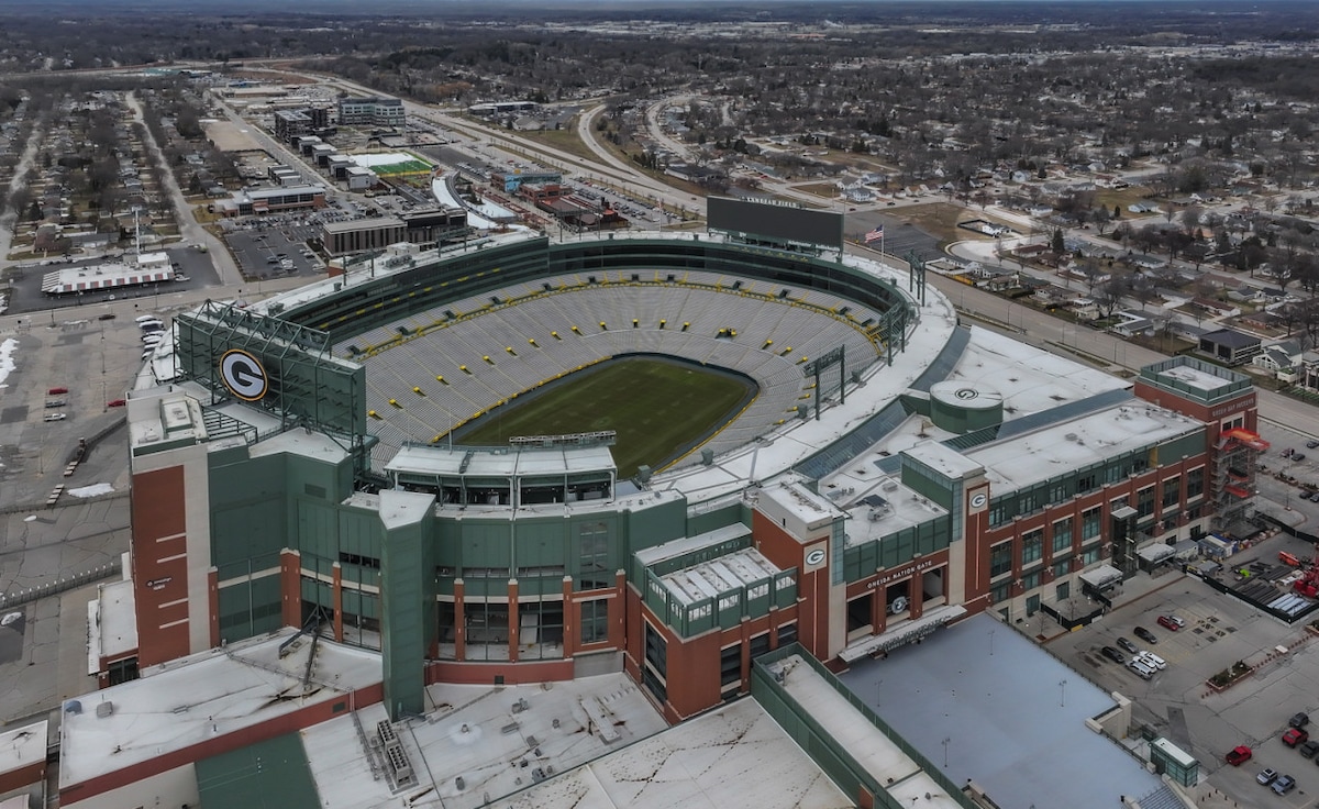 An aerial view of Lambeau Field is presented, showcasing the iconic green and gold architecture. Surrounding landscapes include roads and a variety of structures. The stadium's seating appears organized, with various yellow accents visible throughout the seating sections.