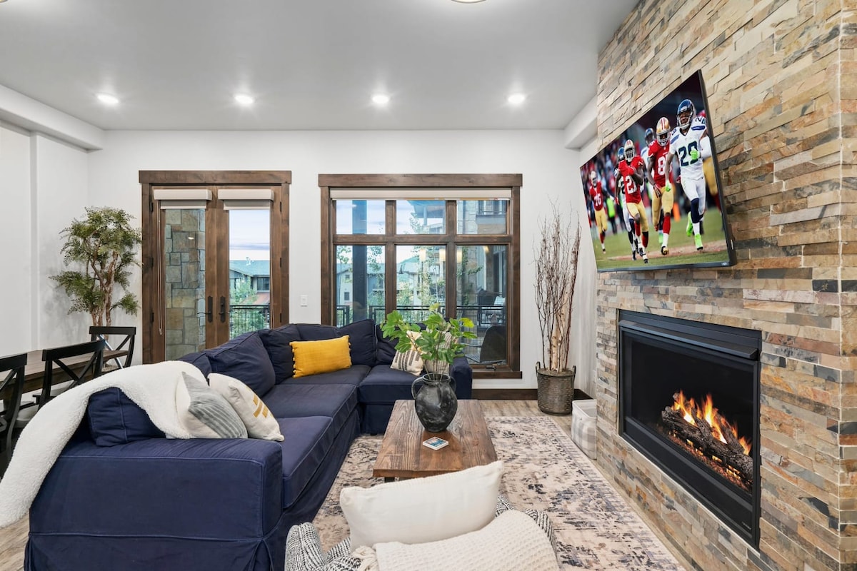 A spacious living room features a cozy sectional sofa positioned near a stone fireplace. A large flat-screen TV is mounted above the fireplace. Natural light comes through multiple windows, enhancing the room's inviting appearance. A wooden coffee table occupies the center, complemented by decorative plants.