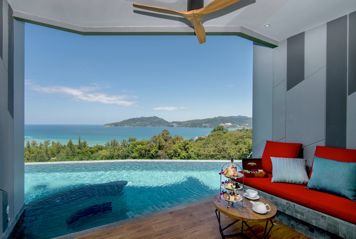 A private balcony features a stunning view of the ocean, complemented by a spacious infinity pool. Comfortable seating with vibrant cushions is arranged nearby, providing a serene space to relax and enjoy refreshments. The natural scenery includes lush greenery and distant hills.