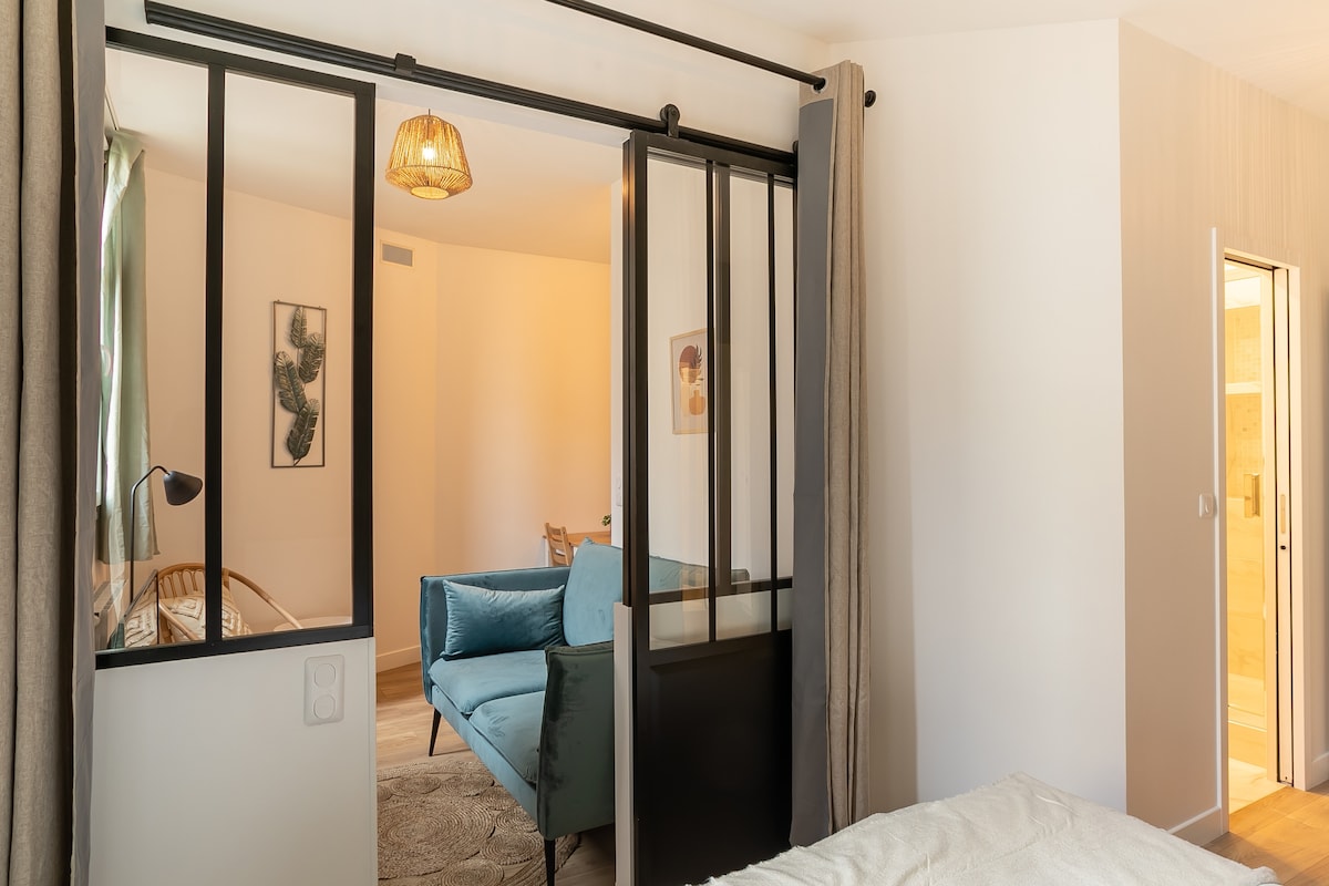 The image shows a divided space featuring a cozy sleeping area with a bed, a sliding glass door, and a view into a stylish living area. A comfortable sofa with soft cushions is visible, situated beside a decorative lamp, welcoming guests into the inviting ambiance.
