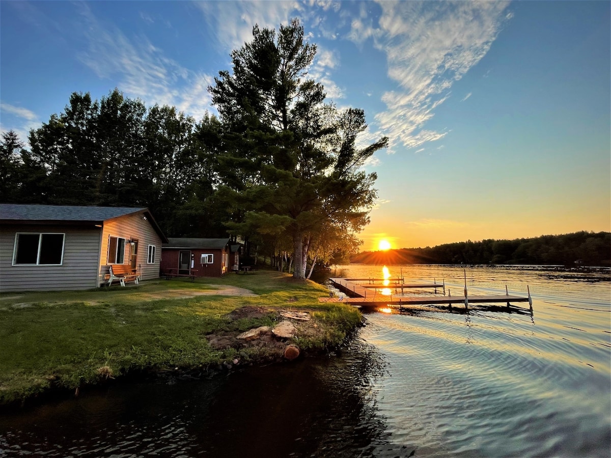 A lakeside view captures the sun setting over Long Lake, with reflections dancing on the water's surface. The well-kept green lawn leads to a dock, providing access to the lake, while trees frame the scene and a cozy cabin is visible nearby.