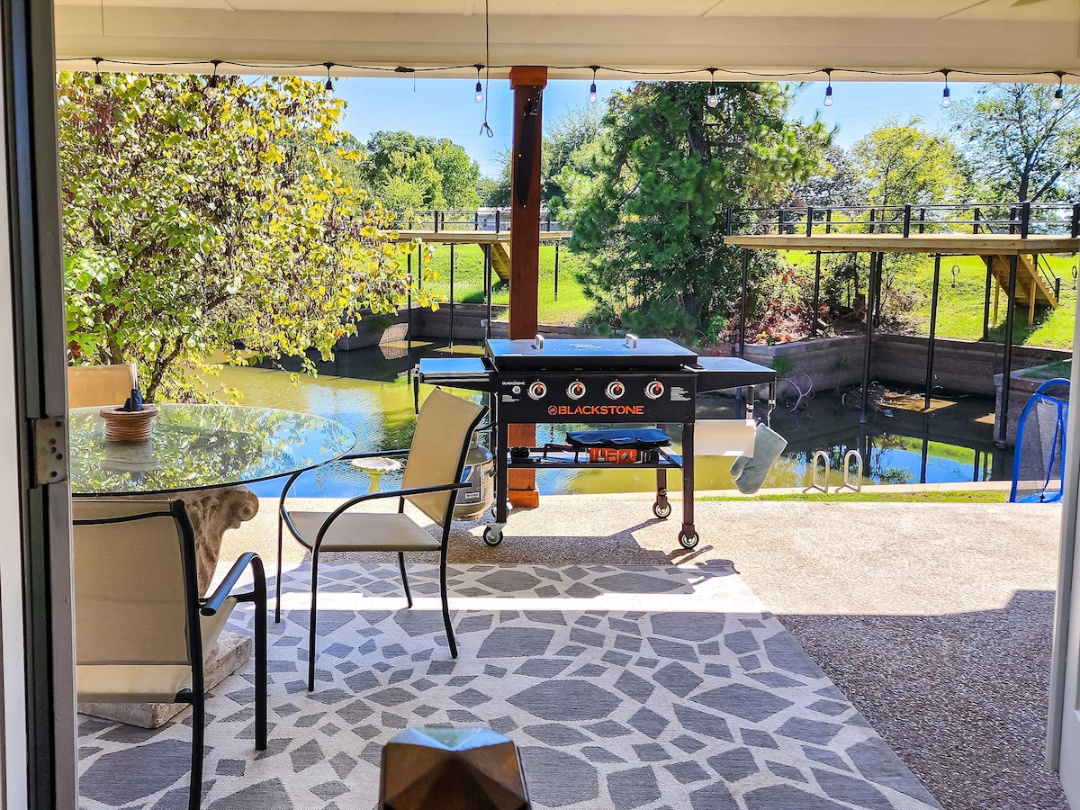 An outdoor dining area features a glass table with two chairs, set against a serene lake view. A gas grill stands nearby, with greenery and a covered boat slip visible in the background. Soft lighting enhances the space, inviting relaxed meals outside.