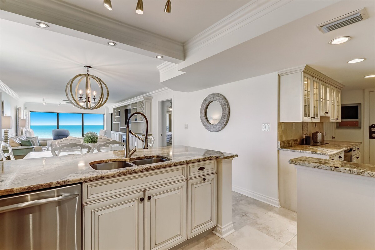 A spacious kitchen area features light cabinetry and granite countertops, with a double sink positioned under modern pendant lighting. The open layout allows for a view of the ocean through large glass windows, connecting the cooking space to the inviting living room beyond.