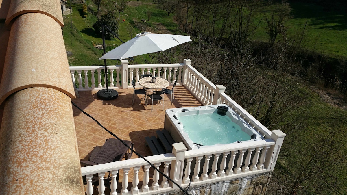 An outdoor terrace is featured with a private jacuzzi and a round dining table shaded by a large umbrella. A lounge chair is positioned nearby, set against a backdrop of lush greenery and open space.