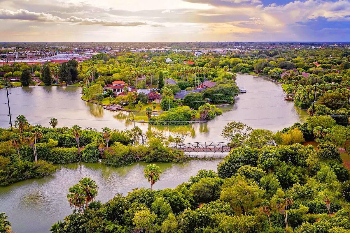 Aerial view of a lush, green landscape featuring winding waterways and dense trees. The scene includes homes nestled among the greenery, with a wooden bridge crossing a waterway. The sky is partly cloudy, casting a warm glow over the serene environment.