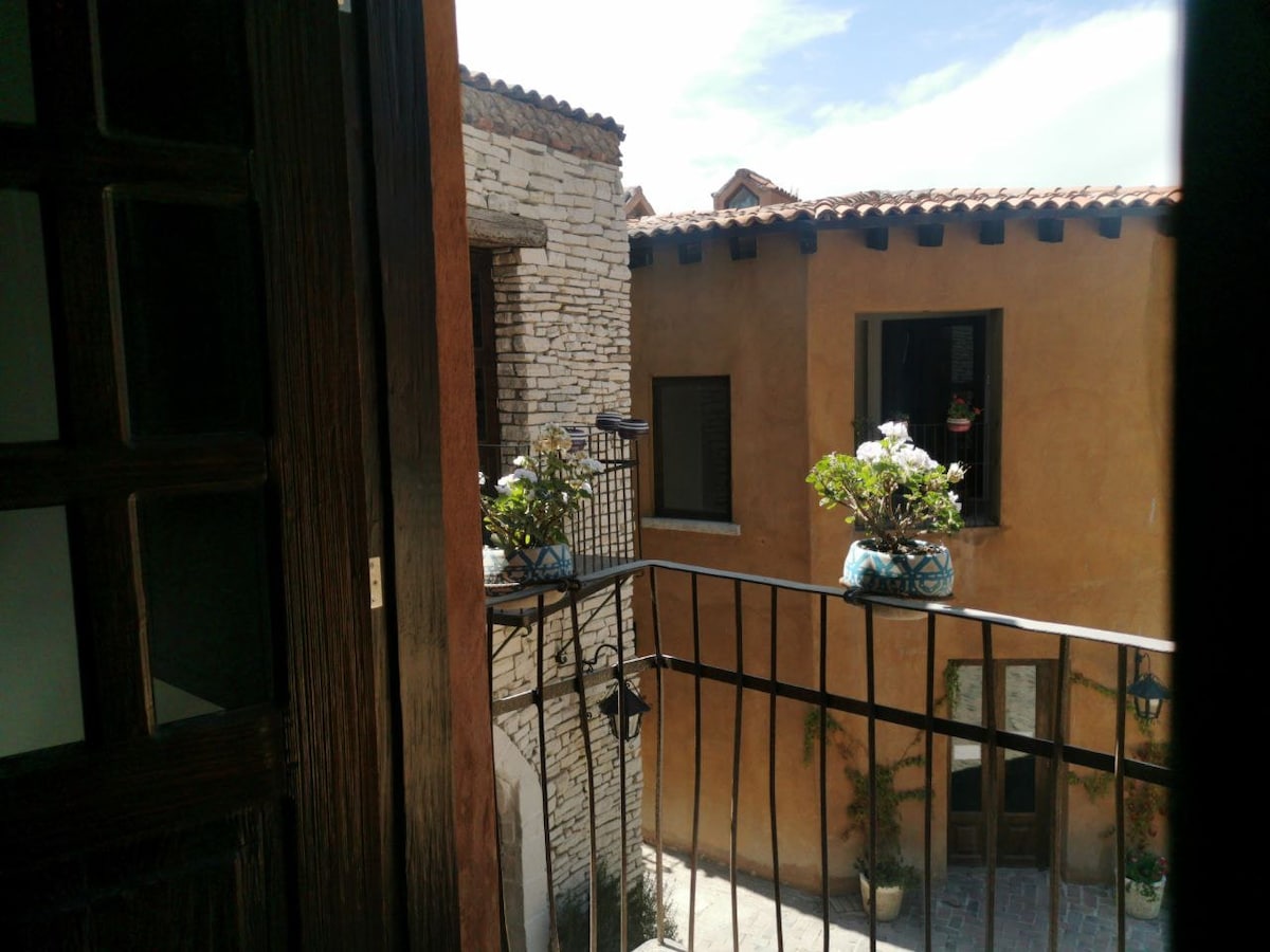 A view from a balcony offers a glimpse of charming architecture featuring warm earth-toned walls and wooden accents. Potted plants with white flowers adorn the balcony railing, providing a touch of greenery against the backdrop of neighboring buildings and a bright blue sky.