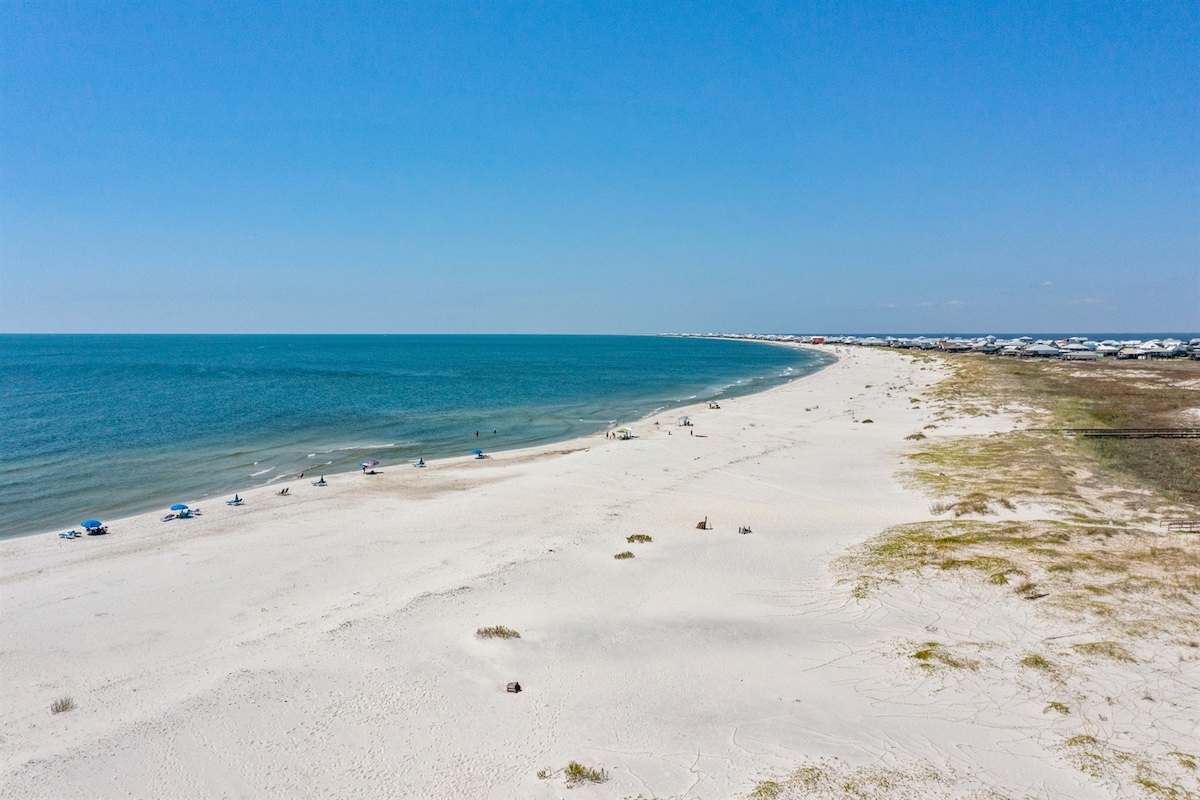 A wide-angle view of a serene beach where soft, white sands meet the calm, blue waters of the Gulf of Mexico. The shoreline extends into the distance, with a few beachgoers relaxing under umbrellas, showcasing an inviting atmosphere for outdoor enjoyment.