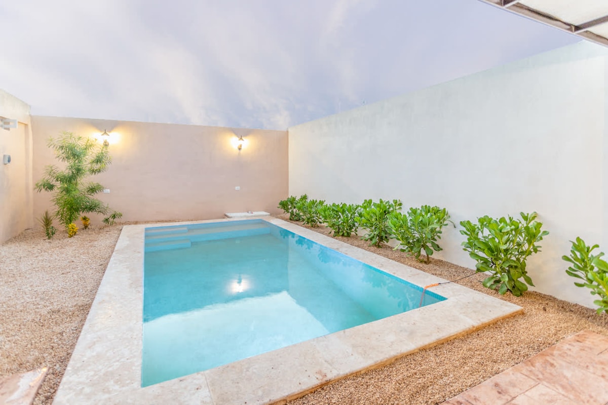 A private swimming pool is surrounded by a smooth stone deck and landscaped with lush green plants. Light fixtures on the walls illuminate the area, creating a serene atmosphere. A soft, warm sky serves as the backdrop, enhancing the tranquil setting.