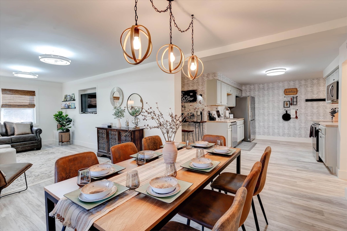 A dining area features a rectangular wooden table set for six, with neutral plates and glassware. Unique pendant lights hang above, and a cozy seating arrangement is visible in the background. The modern kitchen is partially seen, showcasing contemporary cabinetry and appliances.
