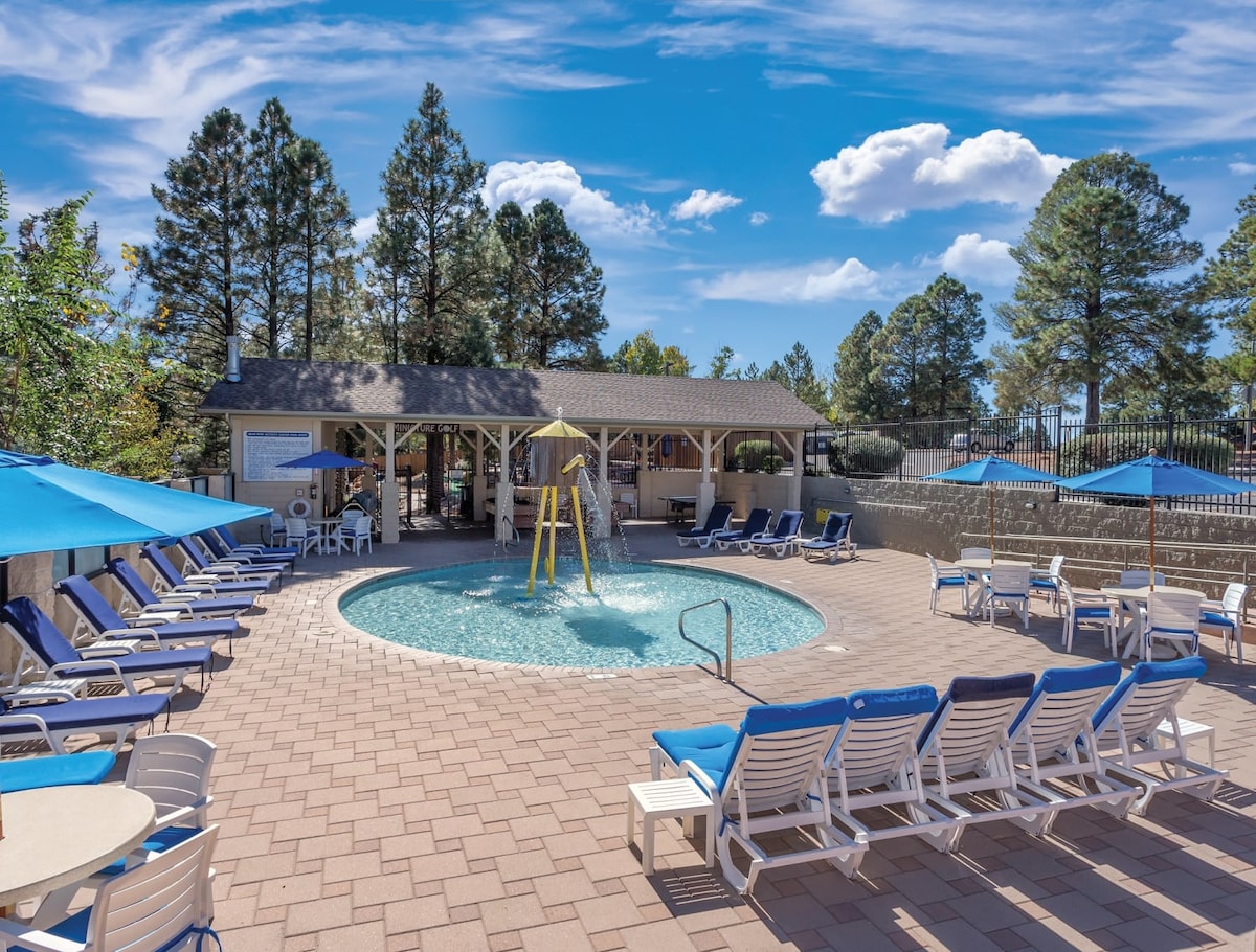 An outdoor pool area is surrounded by lounge chairs under blue umbrellas, providing shaded seating. A tall slide stands beside the pool's water, and a shaded gazebo offers a space for relaxation. Pine trees frame the background against a clear sky.
