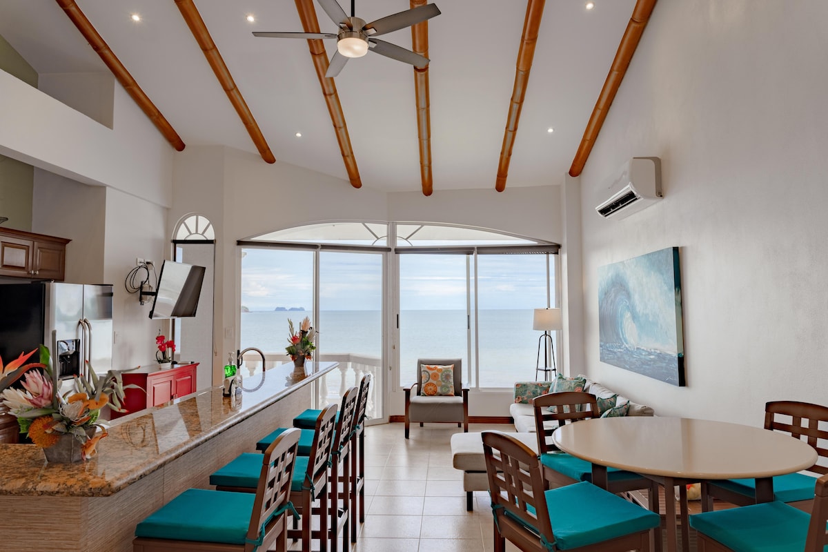 An open-concept living space features vaulted ceilings with wooden beams and large windows showcasing ocean views. A kitchen bar with stools and a round dining table are visible, while comfortable seating arrangements create a welcoming atmosphere. Decorative plants and light artwork add color to the space.